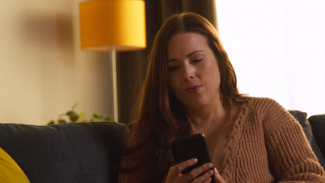 Woman-Sitting-On-Sofa-At-Home-Using-Mobile-Phone-To-Check-Social-Media-Message-And-Read-News-Article-1